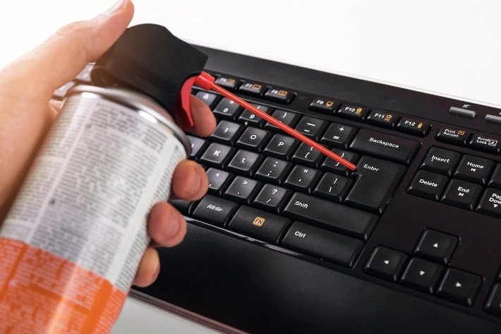 Una imagen que muestra a una persona usando un pulverizador de aire para limpiar el teclado de una computadora portátil.  El teclado está inclinado en ángulo y el pulverizador de aire se dirige hacia las teclas.  Esta imagen muestra un método para reparar las teclas del teclado de una computadora portátil presionadas automáticamente.