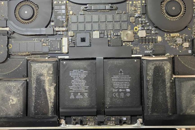 The photo presents a top-down view of the interior of a MacBook A1398, highlighting the motherboard and the battery. The components are covered with a noticeable amount of dust and debris, which is common in devices that haven't been cleaned regularly. The dust accumulation is especially visible on the battery surface, the fan on the left, and the logic board. 