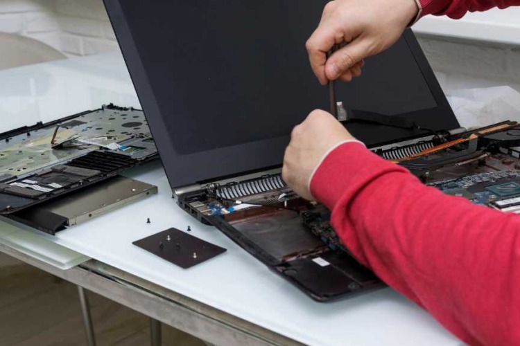 The image depicts a person in the process of disassembling a laptop. The laptop lies open on a white surface, and various components are visible inside. The person's hands are shown using a screwdriver on the laptop's interior, suggesting a step in the maintenance or repair process, possibly related to reapplying thermal paste.